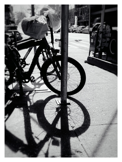 Monochrome photo of what looks like a long dark staff poking vertically through the number 8. It’s actually the aligned silhouette and shadow of the front wheel of a bicycle, backlit by the sun, leaning against a metal post on a city sidewalk.