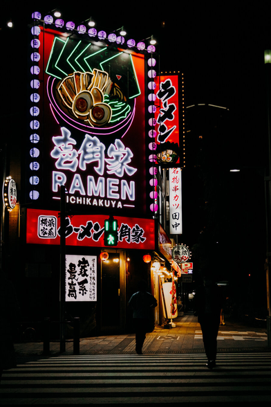 nightly photo of a very large neon sign for a ramen shop across the street, with a person walking over a crosswalk in front of it