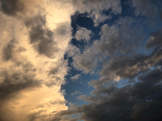 Photograph of an evening blue sky with clouds, with sun light turning to yellow and illuminating white and grey clouds. The left side of the picture is occupied by a bright white and thick cloud formation, with smaller fluffy grey clouds in front. On the right side, fluffy white and grey clouds in deep blue sky.