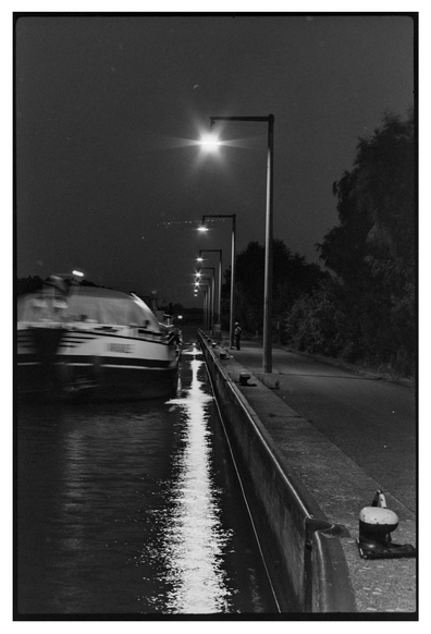 Black and white photograph in portrait orientation. A night scene at a canal. Some lights are reflected in the water producing a strong line of light. A cargo ship is moving fast enough to produce motion blur. Somebody is standing at the shore.