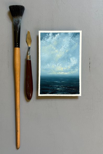 Original seascape oil painting by Tisha Mark, "Hopeful Morning No. 2501" 6"x4" oil on Arches oil paper (2025), shown with a paintbrush and palette knife for scale. Textured seascape painting with white atmospheric clouds touched by yellow sunlight in a blue sky over a deep blue sea. Yellow sunlight from the sky reflects in the water below.