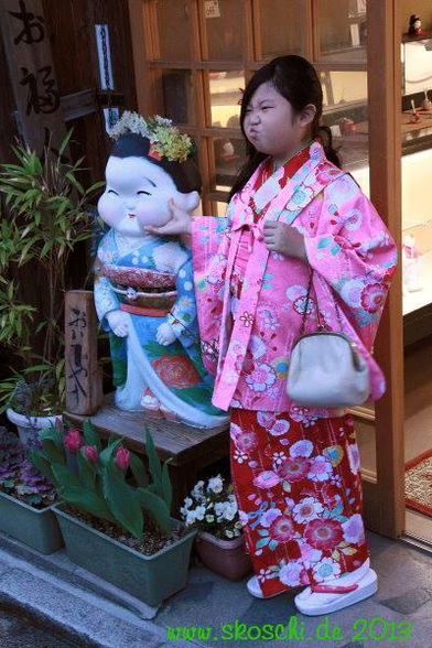 Ein junges asiatisches Mädchen im pinken Kimono, posiert vor einer Statue mit aufgeblähten Wangen, und tut es ihr nach. 