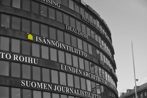 A mainly black and white photograph of the side of a donut-shaped building, with several floors creating circles of windows, stacked. Between the row of windows are company and brand names advertising the name of the companies whose offices are in the building.

There is a bright yellow logo on the left of one of the names which has retained its color, even though the rest of the photograph is black and white. It's a simplified house shape with a dot above on the left, which could be understood as a chimney.