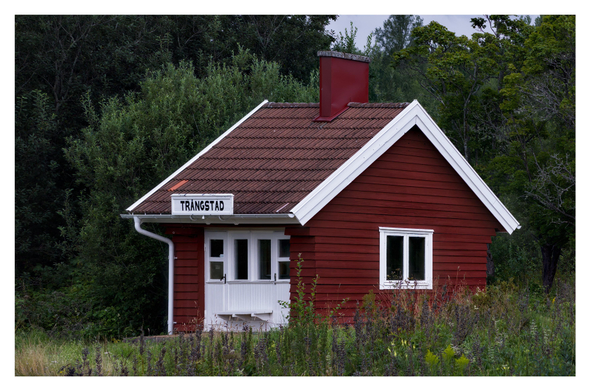 En liggande bild på ett litet rött trähus för tågresenärer. Skylten på taket säger ”TRÅNGSTAD”. Grönska och träd runt omkring.