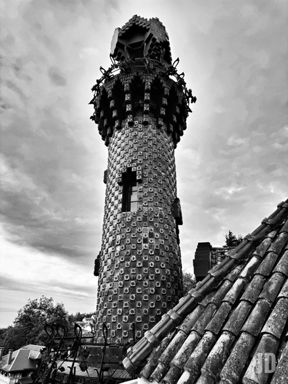 Una imagen en blanco y negro muestra una torre alta y cilíndrica de arquitectura ornamentada, fotografiada desde un ángulo bajo contra un cielo nublado. La torre está densamente cubierta con un patrón repetitivo de pequeñas baldosas cuadradas, cada una con un círculo central, lo que le confiere una textura muy detallada y distintiva. Hacia la mitad de la torre, se distingue una ventana con un marco en forma de cruz. La sección superior es más ancha y elaborada, culminando en un remate de estilo geométrico o cubista con formas angulares. En el primer plano inferior derecho, se ve una porción de un tejado de tejas de arcilla antiguas y desgastadas, con algunas manchas de musgo. Al fondo, a la izquierda, se aprecian algunas copas de árboles y estructuras distantes.