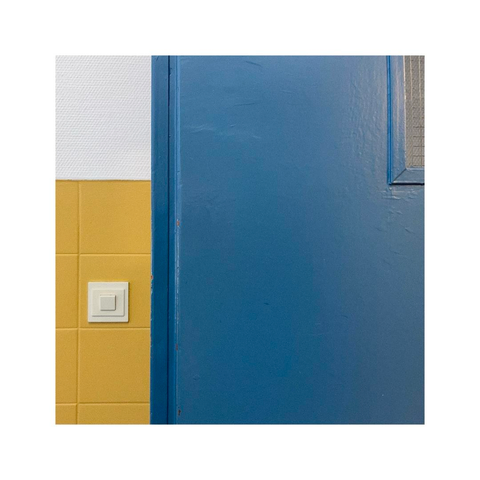 another square frame showing a detail of a dark blue door. the door sits in front of a yellow painted wall and has a little window which is only visible in parts. on the yellow wall sits a white light switch which is as square as the whole frame