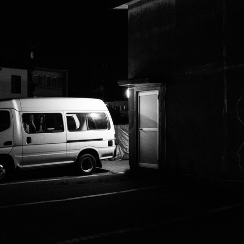 This black-and-white photograph shows a nighttime urban scene featuring a white van parked on a street next to an illuminated storage building or warehouse structure. The building appears to have glass panels or windows with internal lighting that creates a bright rectangular glow against the dark surroundings. Behind these elements, there are residential buildings visible in the shadows. The van has multiple windows and appears to be a commercial or passenger vehicle. The composition captures the contrast between the brightly lit storage structure and the darker urban environment, with the white van serving as an intermediate tonal element. The scene represents typical mixed-use urban infrastructure where residential and commercial/storage facilities coexist.