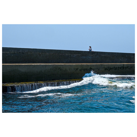 A seawall extending horizontally across the frame. Above the barrier, two persons are walking. The lower portion of the image shows water with waves crashing against the base of the barrier. The peak point of the wave coincides with the position of the two people. The water is a deep blue, with white foam and splashes visible. The sky above is clear and light blue, free of clouds. The overall scene is bright and sunlit. The composition emphasizes the contrast between the solid structure and the fluid movement of the water. (with help of Mistral.ai)