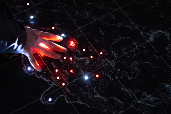 Same map, different perspective (from the top). A man's hand gently touches the map, causing the LEDs to light up. There are about a dozen of them on the map, some of which are under the man's hand. Bright light shines through his fingers.