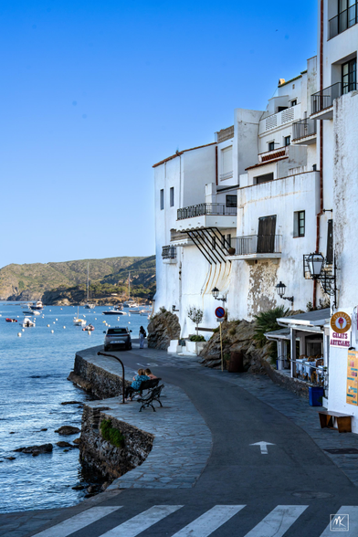 Color photo of white buildings along a road with water on the other side. 