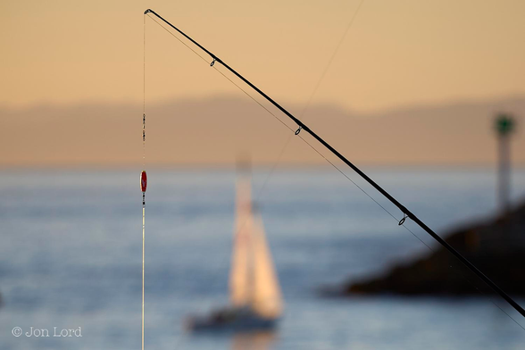 This is a colour sunset photo of a sailing boat, a fishing rod and line. 
In the foreground and stretching from the lower right corner diagonally upwards towards the upper left corner is a fishing rod, sharply in focus. Hanging from the upper end of the rod is the fishing line, the lower part translucent from the light of the setting sun. The fishing rod and line form a 'right-angled triangle' with the image's lower margin forming the third leg. Within this triangle is a sailing boat, its sails glowing from the near set sun: the sails also forming a triangle. The boat is sailing from left to right towards the harbour, a short distance away. In the right is the harbour breakwater, deep in shadow, as it slopes down towards the water forms another triangle. The blue and blurred out of focus sea reaches upward to the halfway up point in the frame. Above is a band of distant mountainous terrain rendered a golden or caramel colour by the sunset. Above is a clear and cloudless sky, exactly the same colour as the terrain but with a lighter tone. 
The location: Redondo Beach, California. The terrain: the Santa Monica Mountains (2013).