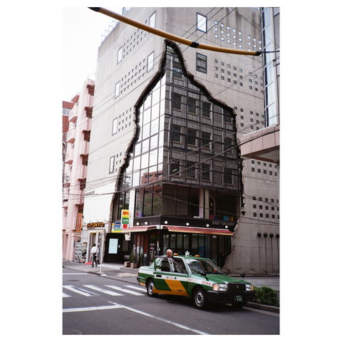 The image shows an office or shop building, with one side appearing to have broken away, revealing a glass and steel structure beneath. In front of the building is a dark green taxi.