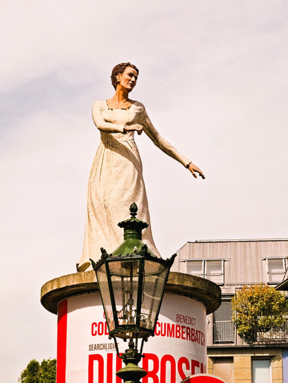 A column saint in the form of a bride in a long white dress. She is looking to the side and moving her arms as if she were dancing. She is standing on an advertising column, in front of which there is a pretty street lamp. The sky behind her is cloudy.