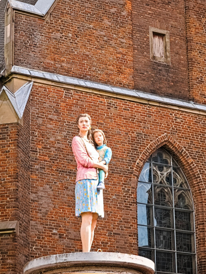 Column saints in front of a church. It is a woman in a short skirt holding a child in her arms. Both are looking seriously into the distance. This sculpture is called 'The stranger'