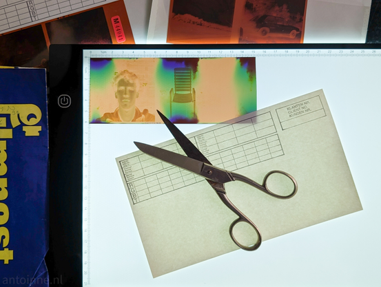 The central focus is a backlit film strip laid across a glowing white light pad with a ruler printed along the top and left sides. The film strip contains two distinct images: a portrait of a person, and a chair. The film has a distinct orange tint, common for negatives.

A pair of metal scissors with a classic design lies diagonally across the light pad, resting on top of a light-green paper form with grids and text. The scissors are positioned to cut the film strip. 

Other photography-related items are scattered around the workspace, including a blue booklet titled "Filmpost" and other film negatives and strips.