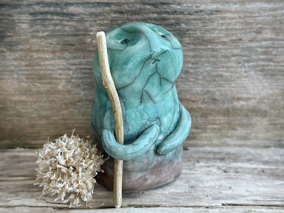 against a background of grey-brown driftwood, a raku ceramic nature spirit warrior sculpture. it is glazed in a gradient from warm brown to forest green (bottom to top) and carries a stick in one of its arms. it has a sad and worried but determined look on its gentle face. next to it on the left is a pale, beige-coloured dried chive flower.