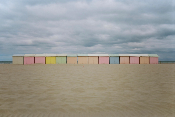 A row of paste-coloured cabins on a sand beach, under a cloudy sky.