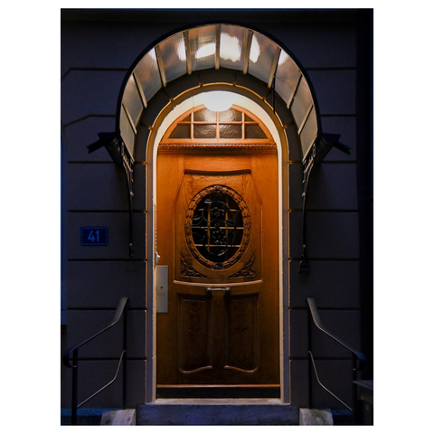 an entrance to a building at night. The focal point is a wooden door with intricate carvings and a decorative oval window featuring a wrought iron design. Above the door is a semi-circular transom window framed by a light fixture that illuminates the entrance. The building number "41" is displayed on a blue plaque to the left of the door. The walls surrounding the door are painted in a dark color, and there are metal railings on either side of the steps leading up to the entrance. (with help of mistral.ai)