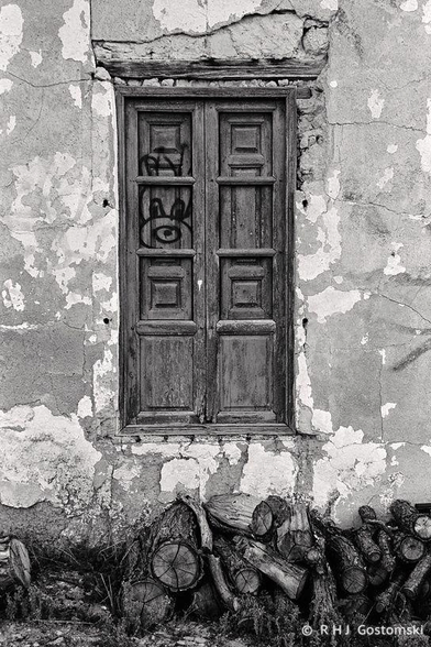A weathered wooden window with graffiti on the wooden shutters is set in a cracked, peeling wall. A pile of cut logs rests beneath the window. Captured on Ilford FP4 with a Nikon FE2
