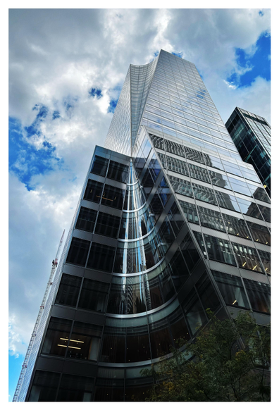 A view of the entire tower, which dwarfs a tree at lower right and a vertical construction crane at its left edge as it rises into a cloudy sky. We see more clearly how the concave curvature of the tower’s bottom cone reflects elongated images of the skyscrapers behind the camera, seeming to converge at the cone’s apex.