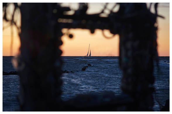 Farbfoto im Querformat mit weißem Rand. Zu sehen ist ein unscharfer Fensterrahmen aus Treibholz, wenn man hindurchschaut, sieht man am Horizont ein Segelschiff vor orange-gelbem Himmel.