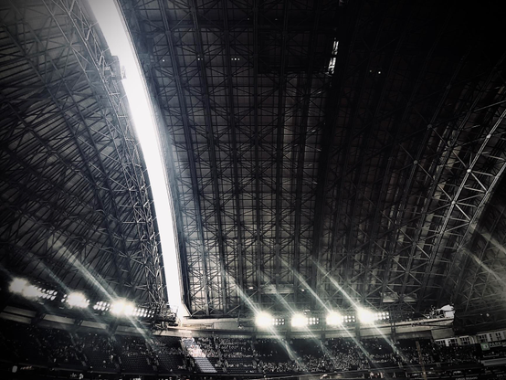 Photograph of the roof of a baseball stadium just starting to open before the game, taken from inside, showing only a crack at that moment and sunlight coming in. The metal structure of the roof is visible, as well as the bright spotlights just above the stands, starting to fill with people. The grainy texture of the picture taken with a phone makes it look like a drawing. The is Rogers Stadium, the home of the Toronto Blue Jays.