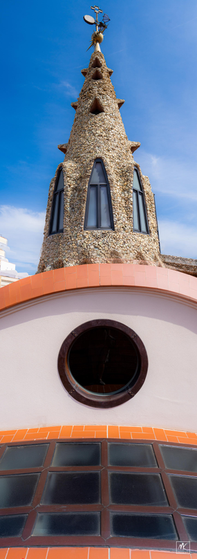 Color photo of a stone-covered rooftop spire with arched windows in it above a round window that is above a glass brick roof surface in the foreground. 