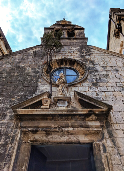 A low-angle photograph shows the weathered stone facade of a church building, taken from directly below the entrance in Dubrovnik, Croatia. Above the doorway, a decorative stone pediment frames the base of the facade. Above the pediment is a statue of St Joseph holding a child, standing in a niche. To the left of the statue, a small tree grows directly out of a crack in the wall. Above the statue, a large, ornate circular window is visible, and above that is the church's bell tower, topped with a cross. The sky is blue with scattered clouds.