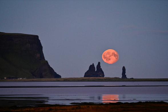A full moon rises above the ocean and the jagged basalt pillars of a sea stack just off shore. The moon’s reflection appears in the still water of the middle distance.