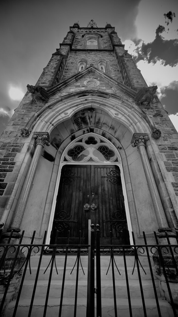Entrance doors to a church, behind a gate and up a short flight of stairs.  Window, door surround and tower have some decorative architectural elements. Black and white picture taken at an oblique upwards angle, and the building seems tall and imposing.