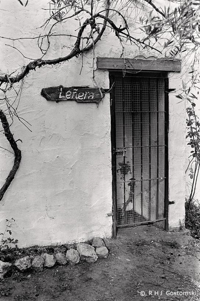 Black and white photo of a rustic building with a wooden “Leñera” sign next to a barred door, shot on Ilford FP4 film. The rough, whitewashed wall and foliage give the photo a timeless rustic look.
