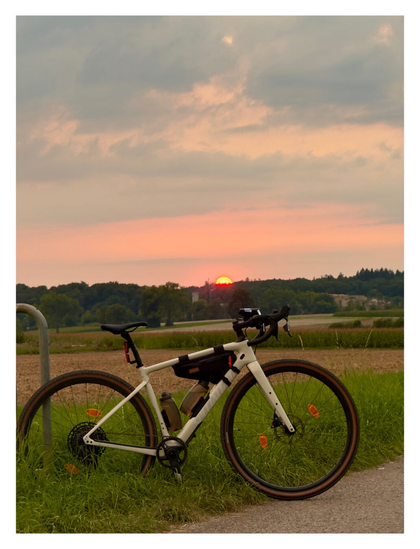 Foto im Hochformat. Ein cremefarbenes Gravelbike steht am Rande eines asphaltierten Weges auf einer Wiese. Dahinter sind Felder, am Horizont geht gerade die Sonne unter und schaut gerade noch so hinter dem Wald hervor. Der Himmel ist orange und blau.