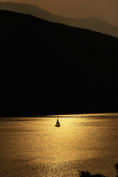 Ein Segelboot auf einem See im goldenen Abendlicht. Im Hintergrund dunkle Berge.