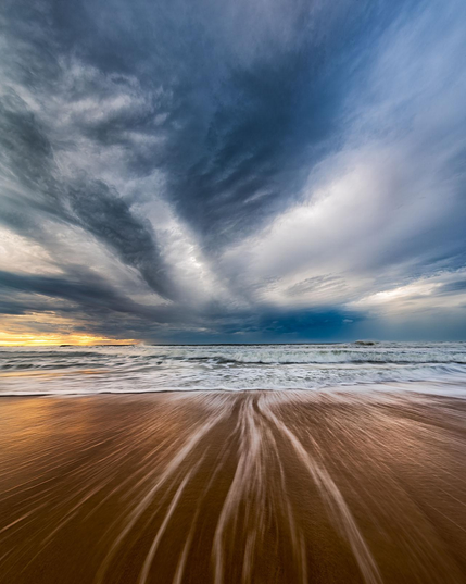 vertical sunrise seascape. The bottom part is brown sand with light grey water streaks pointing towards the center. The sky is dramatic and stormy with menacing clouds loosely resembling a huge bird mid flight. Left side at the horizon is lit with yellow-orange sunrise.