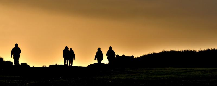 C'est une image en longueur. En contrejour, sur un sol accidenté noir et sans aucun détail ondulant  à peu près à mi hauteur de la photo,  se détachent en noir également sur un ciel dans des tons orangés, cinq  petites silhouettes de personnages hommes et femmes. Une silhouette se tient sur la gauche de l'image, deux très rapprochées, à peu près au deux tiers et les deux dernières sont au centre.