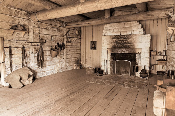 A sepia toned (brown colors like and old time photo) landscape photo of the inside of a large room at a historic trading post. The walls are made of large light colored bricks. The back wall has a large fireplace made of the same brick in the center and the walls on either side are covered in vertical wood plank siding. The fireplace is well used with dark soot stains on the outside. Rough iron implements hang on either side of the fireplace opening. There are flat hearth stones, that are original to the building in front of the fireplace. The flooring is made of wide wood planks. Period equipment such as sacks, baskets, hand wrought iron tools hang from the walls. A painting of a man in a top hat hangs on the wall on the left side of the fireplace.