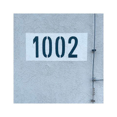 a square image of a number sign printed on the corner of a wall. the wall is painted in a very light gray, the number sign has a white background. the sign is showing the number 1002, the numbers are printed in black. there's a lightning arrester running down the wall on the right side of the number sign