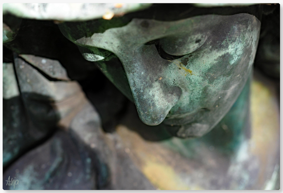 Porträt einer weiblichen Grabfigur von oben. Der Fokus liegt auf den geschlossenen Augen und der Nase. Patina hat die Figur in viele unterschiedliche Grün-und Brauntöne gefärbt. 
Portrait of a female funerary figure from above. The focus is on the closed eyes and nose. Patina has coloured the figure in many different shades of green and brown.