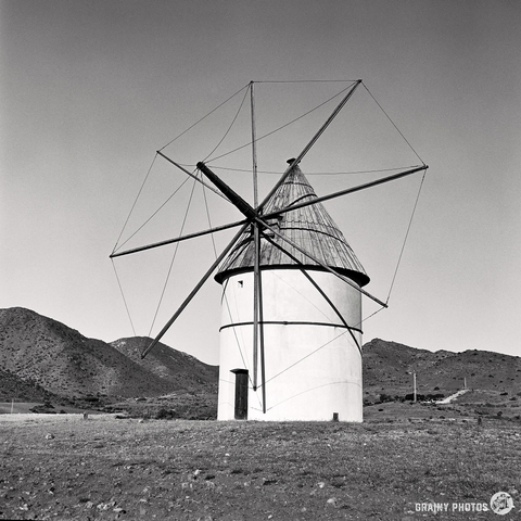A black and white image, captured on Kentmere 100 film with a Mamiya C220, features a traditional restored windmill with a conical roof and slender blades. It stands on a stony landscape with hills in the background under a clear sky.