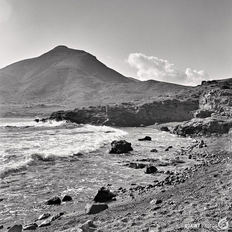 A black and white photograph, shot on Kentmere 100 with a Mamiya C220, depicts a rocky shoreline with waves gently crashing against it. A mountain looms in the background under a mostly clear sky, adding depth to the serene coastal landscape.