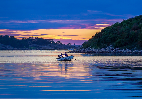 Ein kleines Ruderboot mir 2 Personen in einer Meeresbucht bei Sonnenuntergang. Das Boot in in der Mitte des Bildes. Im Hintergrund ragen zunächst von rechts und dahinter von links bewachsene Felshügel in das Wasser. Um das boot kräuselt sich das Wasser, die Oberfläche wirkt fast weiß. Vor dem Boot glitzert das Wasser wie ein Schein fast im Halbkreis im orange der untergehenden Sonne. Die Schatten des Boots und der Ruderer spiegeln sich darin. 
Das Orange des Sonnenuntergangs sieht man nur zwischen den Felsketten. Ein kleiner leuchtender Schein lässt erahnen, wo die Sonne gerade hinter den Wolken verscwunden ist. Auch der Himmel am oberen Bildrand ist eine einzige Wolkendecke, die überwiegend leicht lila erscheint.