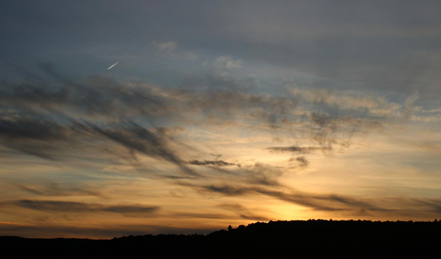 Photograph of a sunset in a blue but cloudy sky, with a yellow and orange light coming from the sun just under the horizon. Grey and white clouds appear to be far away. Other grey and more wispy clouds are "closer" or maybe lower in the sky, and sun rays are filtering through them. In the top left corner, a plane is leaving a bright and white contrail behind.