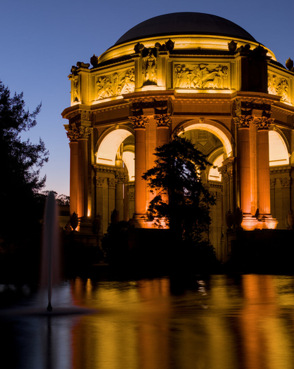 A night time shot of the Palace of Fine Arts in San Francisco. The building is lit up, and reflected in the dark water