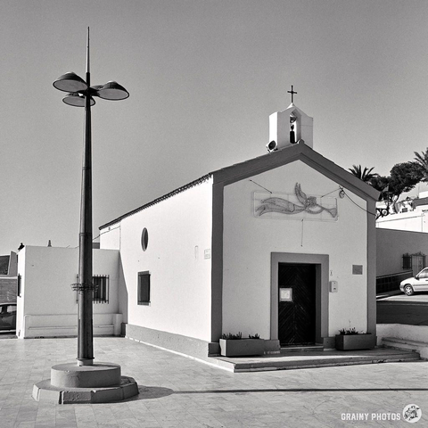 A small, white chapel with a single bell tower stands on a paved area. A modern streetlamp is in the foreground. The chapel door is dark, adorned with not yet removed Christmas decorations above it under the clear sky, while nearby houses are partially visible.