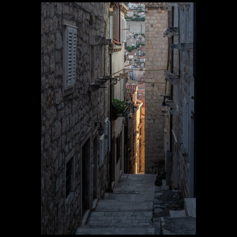 Narrow stone alley with stairs, opening to houses with red-tiled roofs in the distance.