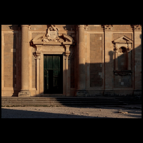 Stone facade of an old building with a large dark door, columns, and steps lit by the morning sun.