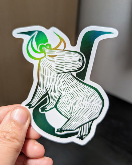 Photo of a left hand holding a diecut sticker. The sticker is a holographic drawing of a capybara Taurus zodiac sign.