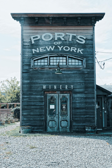 A tall (relative to it's width) building with faded wood siding made of thin boards has elaborate details on the edge of a flat roof and doors that look like they came out of cowboy movies with a single light above it hanging from below a set of three windows with a flat bottom and curved top,  each divided into many panes,  matching the curve of the windows are the words PORTS of NEW YORK and the woed WINERY is above the doors which have the street number 163 on them.  The building is connected to power on the right and is surrounded by a gravel parking lot.