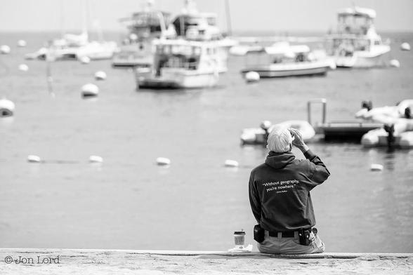 This is a black and white street photo of a man drinking coffee by the sea. 
To the right of and below centre is a senior man sitting on a wall by the waterfront and facing away from the camera. He has a full head of groomed white hair, is wearing a dark, hooded fleece with the words: "Without Geography You're Nowhere." His right arm is raised as he drinks, possibly coffee. His trousers (pants) are fastened with a dark belt with various accessories attached to it. A short distance ahead of our man are the still waters of a bay with a large number of buoys and moored boats. The water extends upward to just short of the upper margin where a bright overcast sky starts.
The location: Santa Catalina, California (2014).