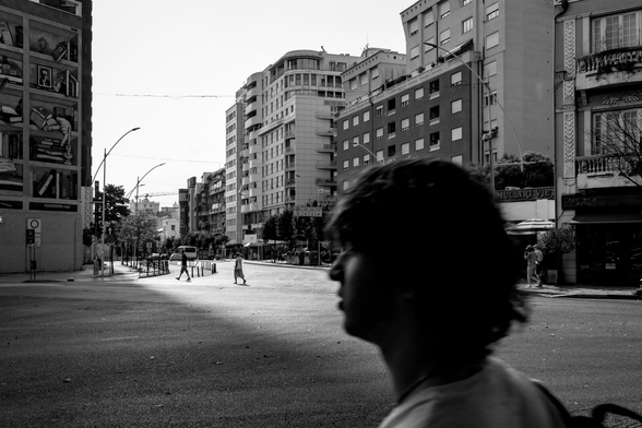 Black and white photo of a city street with tall buildings, pedestrians crossing in the distance, and the blurred silhouette of a person in the foreground.